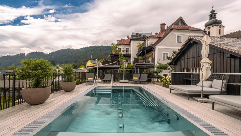 Refugium Lunz, © Niederösterreich Werbung / Maximilian Pawlikowsky A modern pool on a terrace with a view of the surrounding houses and wooded hills in the background.