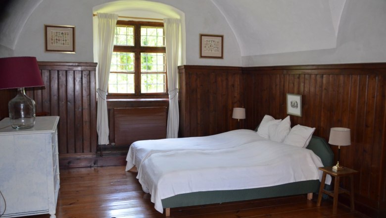 Vacation apartment Vogtei bedroom, © Renaissanceschloss Greillenstein Bedroom with wooden walls, a double bed, a window with curtains and a chest of drawers with lamp.
