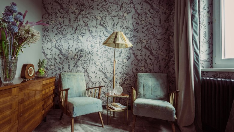 Room, © Matthias Kronfuss A stylishly furnished room with two blue armchairs, a floor lamp and a chest of drawers with flowers and a clock.