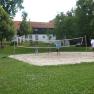 Theater and vacation village Königsleitn, © Theater- und Feriendorf Königsleitn People playing volleyball on an outdoor sand court, surrounded by trees and a building in the background.
