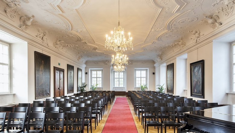 Stift Klosterneuburg, St. Augustine Hall, © Stift Klosterneuburg Elegant hall with stucco ceiling, chandeliers, rows of chairs and red carpet.