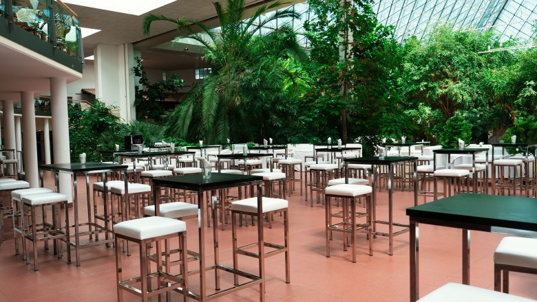 Eventhotel Pyramide, © Viktor Becker Interior view of a modern event room with high tables and chairs, surrounded by lush plants and a glass roof.
