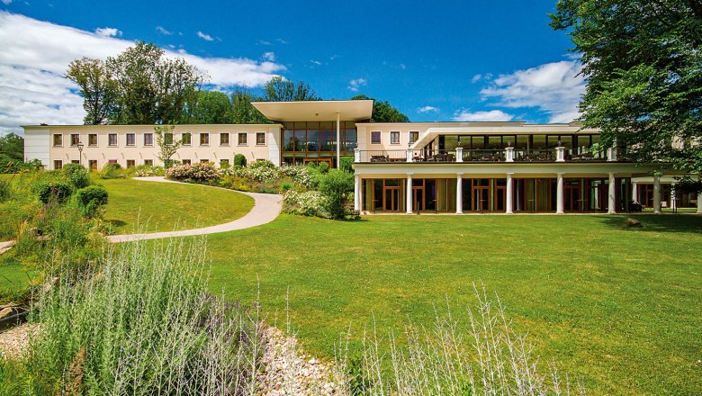 Panorama view, © Schlosspark/Thomas Jandl A modern building with large windows and a terrace, surrounded by a well-tended garden and trees under a blue sky.