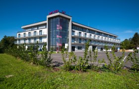 Modern three-storey motel with glass facade and parking lot, surrounded by green landscape.