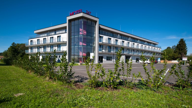 Modern three-storey motel with glass facade and parking lot, surrounded by green landscape.