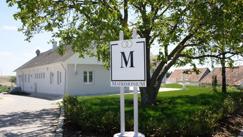 A white building with a "Matrimonium" sign in the foreground, surrounded by a green landscape.