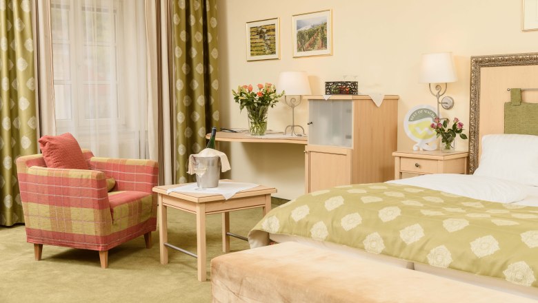 Cozy hotel room with green carpet, red armchair, table with champagne cooler, flowers and bed with green throw.