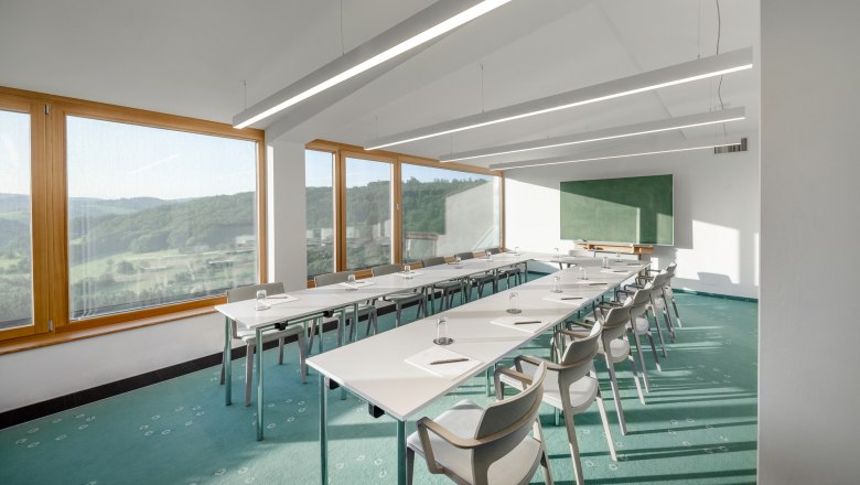 A bright seminar room with large windows offering a view of a green landscape. White tables and chairs are arranged in a U-shape.