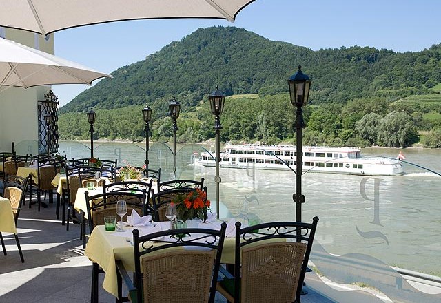 Terrace with covered tables, view of the river and passing ship, surrounded by green hills.
