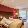 Modern hotel room with double bed, red wall, large window and seating area.