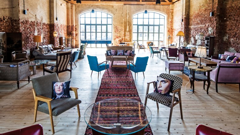 Interior view of a stylishly furnished barn with wooden floor, brick walls and large windows. Various pieces of seating furniture and colorful cushions can be seen.