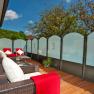 A terrace with rattan furniture, red blankets and cushions, surrounded by glass walls and trees in the background.