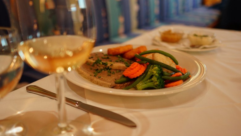 A plate of meat, vegetables and potatoes on a table, with a glass of white wine next to it.
