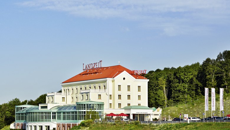 Landzeit Steinh&auml;usl building with red roof and glass facade, surrounded by trees and blue sky.