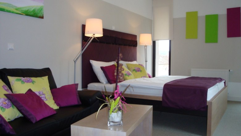Modernly furnished hotel room with double bed, sofa and decoration in shades of purple and green.