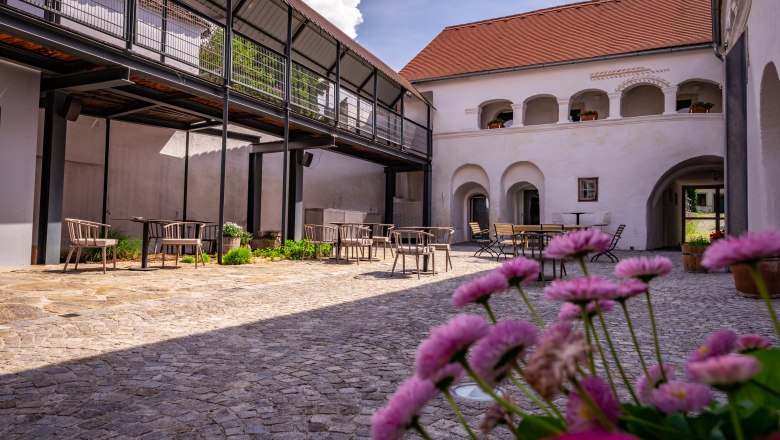 An inner courtyard with cobblestones, tables and chairs, surrounded by buildings with arcades and a modern balcony.