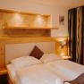 A cozy hotel room with a double bed, light-colored wood paneling, a wooden room divider, and a curtain. There are electrical outlets and light switches next to the bed. Above the bed is a wall niche that is illuminated and painted in an earthy gold tone.
