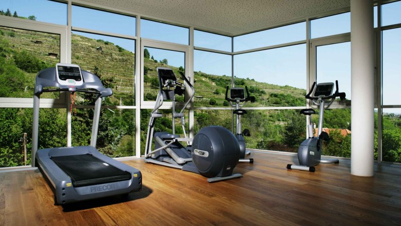 Fitness room with treadmill, cross trainer and exercise bike in front of large windows with a view of green hills.