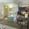 Luxurious hotel room with glass bath, cozy armchairs, table with champagne and fruit, and a large bed with rose petals.