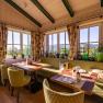 Cozy dining area with large windows, green upholstered furniture and plants, with a view of the countryside.