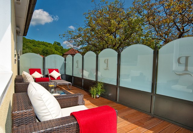 A terrace with rattan furniture, red blankets and cushions, surrounded by glass walls and trees in the background.