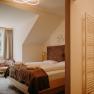 A cozy hotel room with a double bed made of light-colored wood, a window with curtains, and an armchair. In the foreground, you can see into the bathroom. The room has a sloped ceiling on the side with the window.