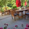 A cozy outdoor dining area with wooden furniture and red umbrellas, surrounded by green trees and flowers.
