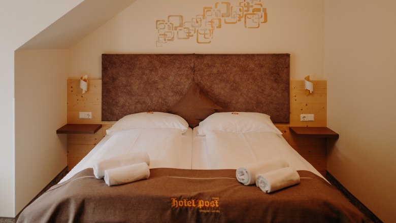 A cozy hotel room with a double bed, featuring light wood paneling and earthy tones.