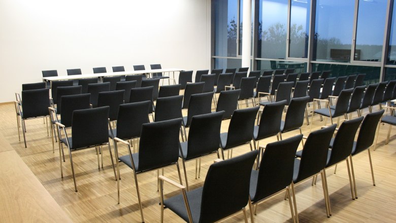 An empty seminar room with several rows of black chairs and a table at the front, large windows at the side.
