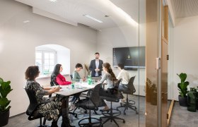 A modern conference room with six people sitting at a table listening to a speaker. A screen in the background.