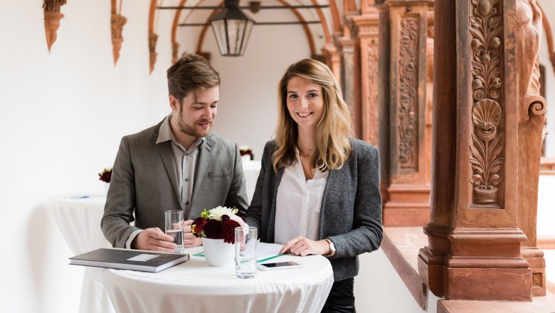 Two people are standing at a high table in a historic building with ornate columns.