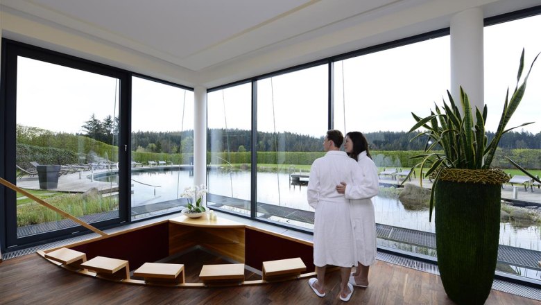 A couple in bathrobes stand in a modern wellness area with a view of a pond.
