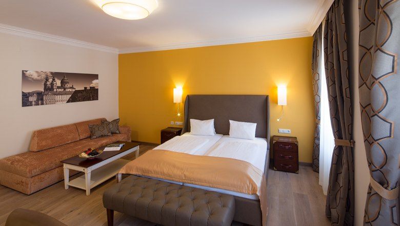 Hotel room with double bed, yellow wall, sofa, table and curtains.