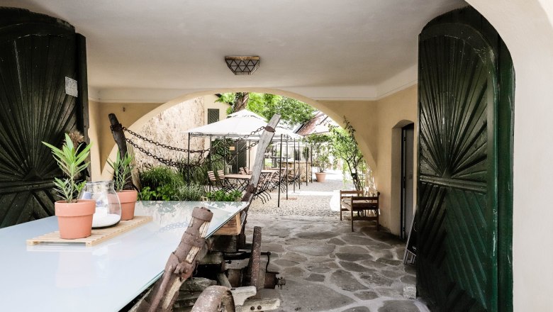A covered passageway with a wooden table and plants leads to a paved courtyard with tables and chairs.