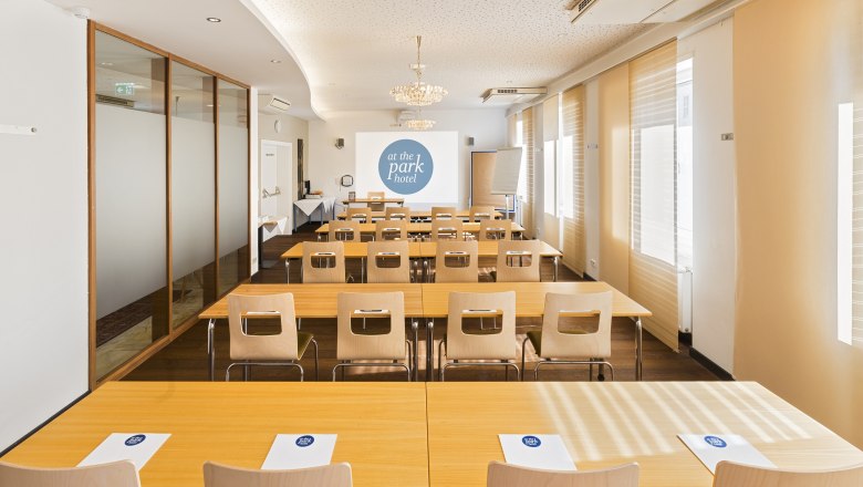 A conference room with tables and chairs, a projector screen and notepads with the "at the park hotel" logo.