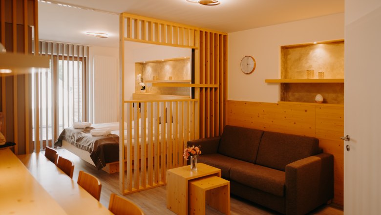 A modern hotel apartment with a double bed, light-colored wood furniture, a pull-out couch, and a wooden room divider. Part of the wall is paneled with wood, and there are niches in the wall painted in an earthy gold.