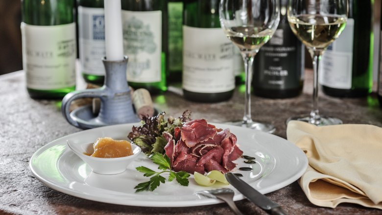 A plate of ham, salad and chutney, surrounded by bottles of wine and glasses on a table.