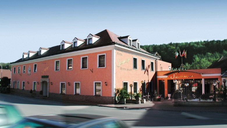 A pink-colored hotel building with several windows and an entrance with a canopy. A car drives past in the foreground.