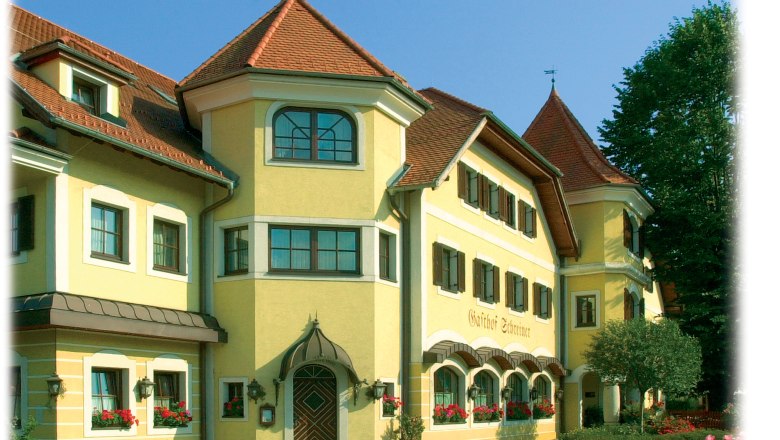 Yellow building with red roofs and many windows, surrounded by trees.