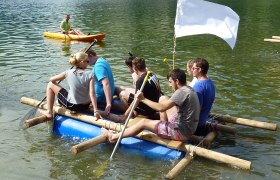 Group of people on a self-built raft on a lake, with a white flag and a kayaker in the background.