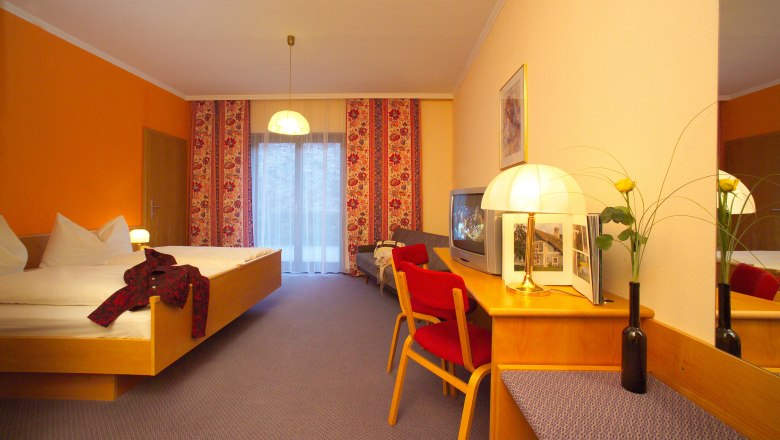 Hotel room with double bed, red chair, TV and lamp on a table. Yellow wall and curtains with a floral pattern.