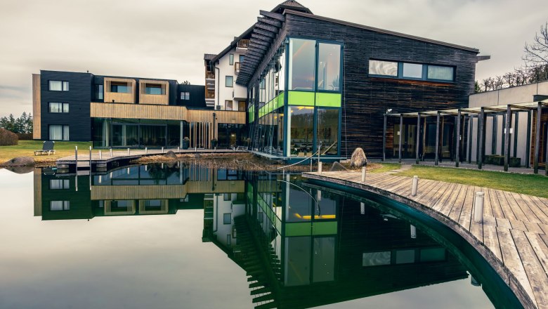 Modern wellness hotel with wooden fa&ccedil;ade and large pond in the foreground.
