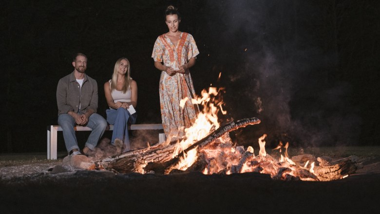 Three people sit and stand around a campfire at night.