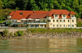 A yellow building with red roofs on the riverbank, surrounded by trees.
