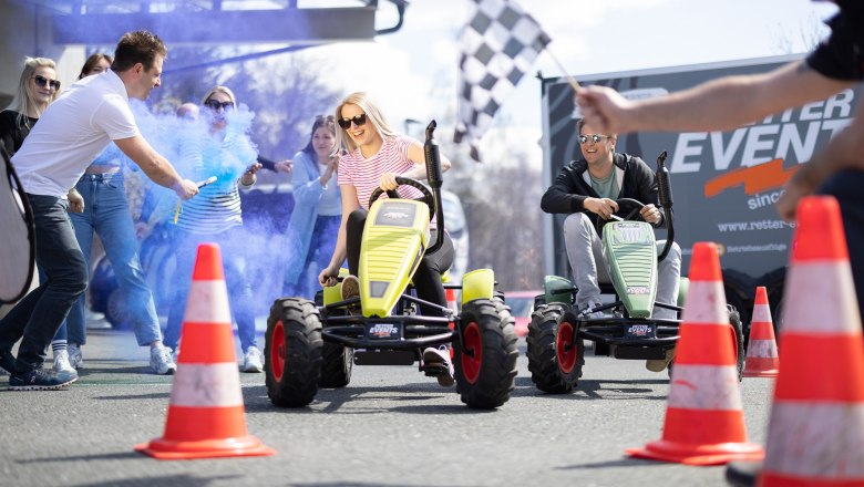 Participants ride pedal tractors at an event with blue smoke and spectators.