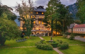 Illuminated building in a garden at dusk, surrounded by trees and mountains.