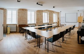 A bright seminar room with a U-shaped table arrangement, chairs, projection screen and flipchart.