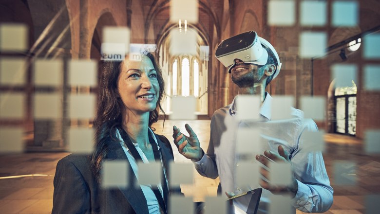 Two people in a historic building, one wearing VR glasses, the other smiling. Blurred squares can be seen in the foreground.