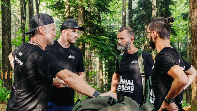 Four men in black T-shirts are standing in the forest and talking.