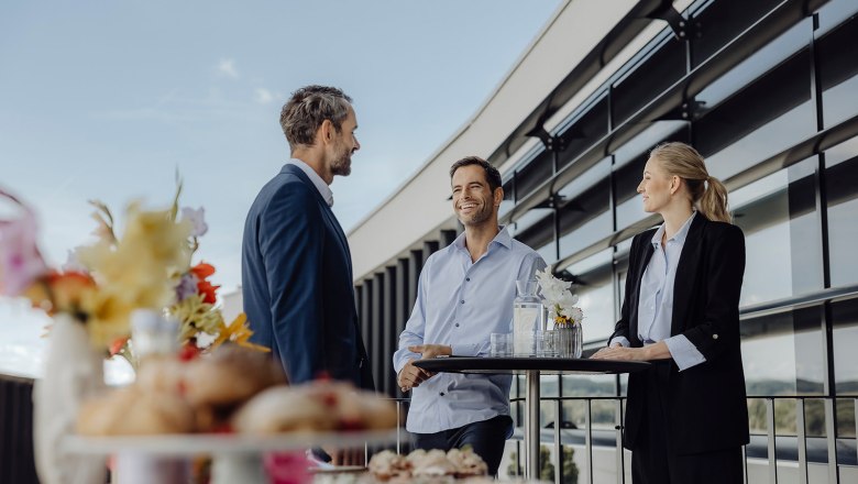Three people are chatting on a terrace next to a modern building.